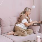 Elderly woman sitting comfortably using a laptop on a pink couch at home.