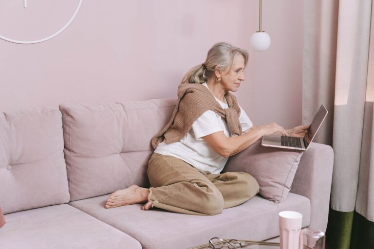 Elderly woman sitting comfortably using a laptop on a pink couch at home.